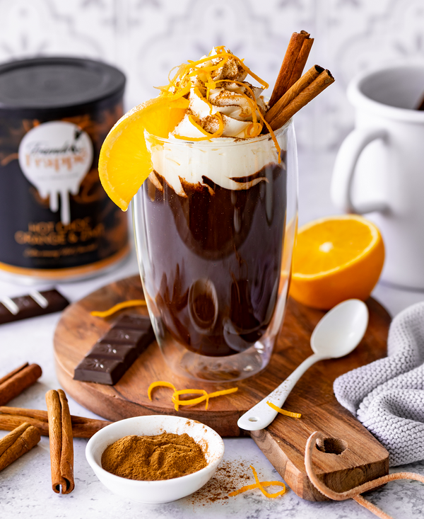 Hot Choc Orange & Zimt, dekorativ angerichtet im Glas mit Sahnehaube, Orangenzesten und Zimtstangen